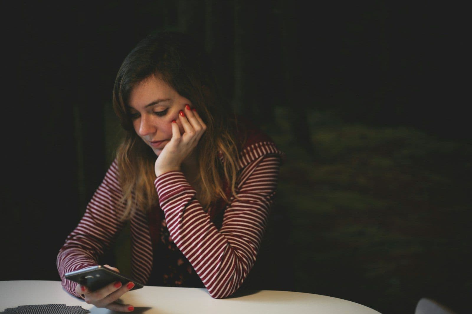 A woman rests her chin on her hand and looks hesitantly at the phone on the table in front of her