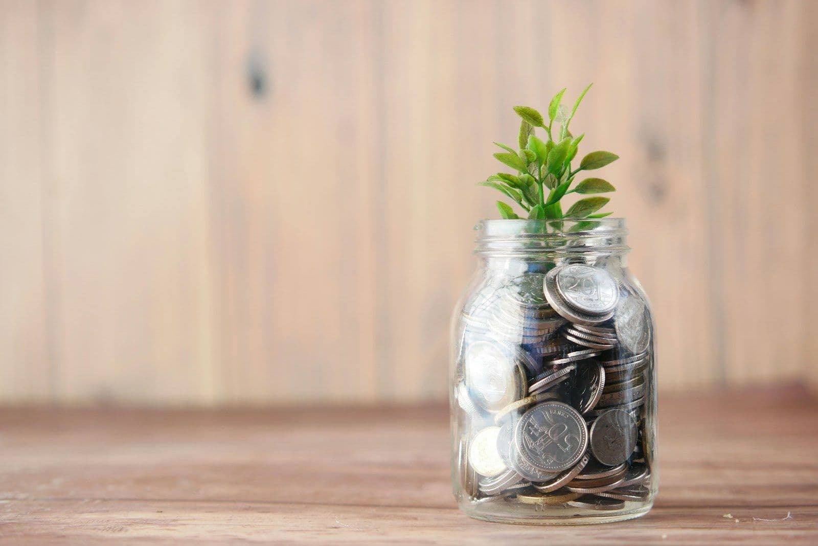 A glass jar of coins with a small green plant growing out of it, on a wooden table