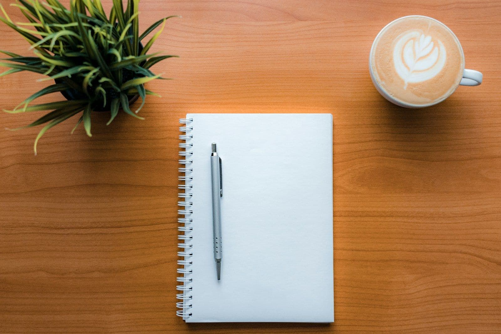 A blank spiral notebook and pen on a wooden desk beside a latte and a small plant